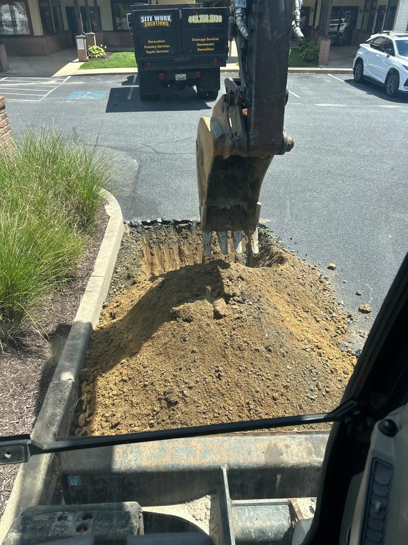 Excavator digging out a section of a commercial parking lot next to a curb, Site Work Solutions dump truck parked behind