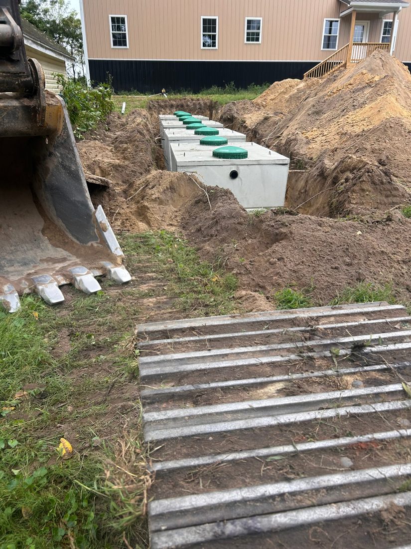 New concrete septic tanks with green riser lids set in an excavation beside a house, excavator bucket and tracks in the foreground