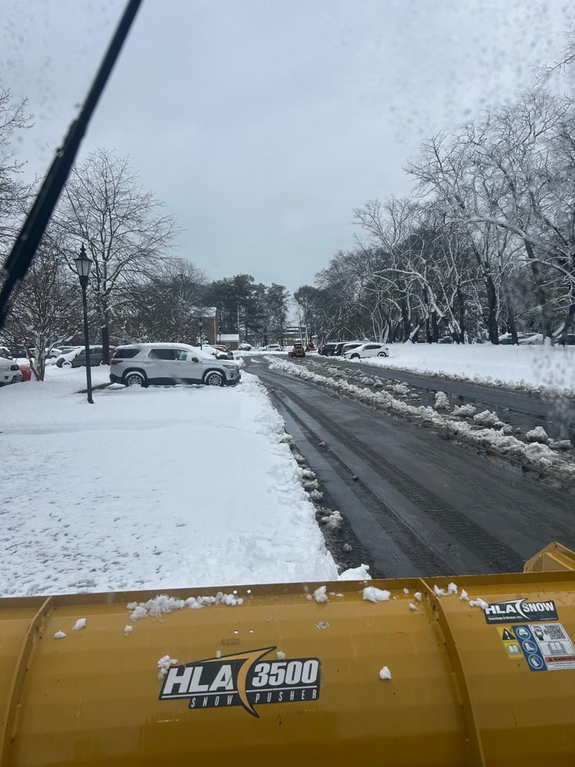 View from the cab over a yellow snow pusher, clearing a snow-covered commercial parking lot road lined with parked cars